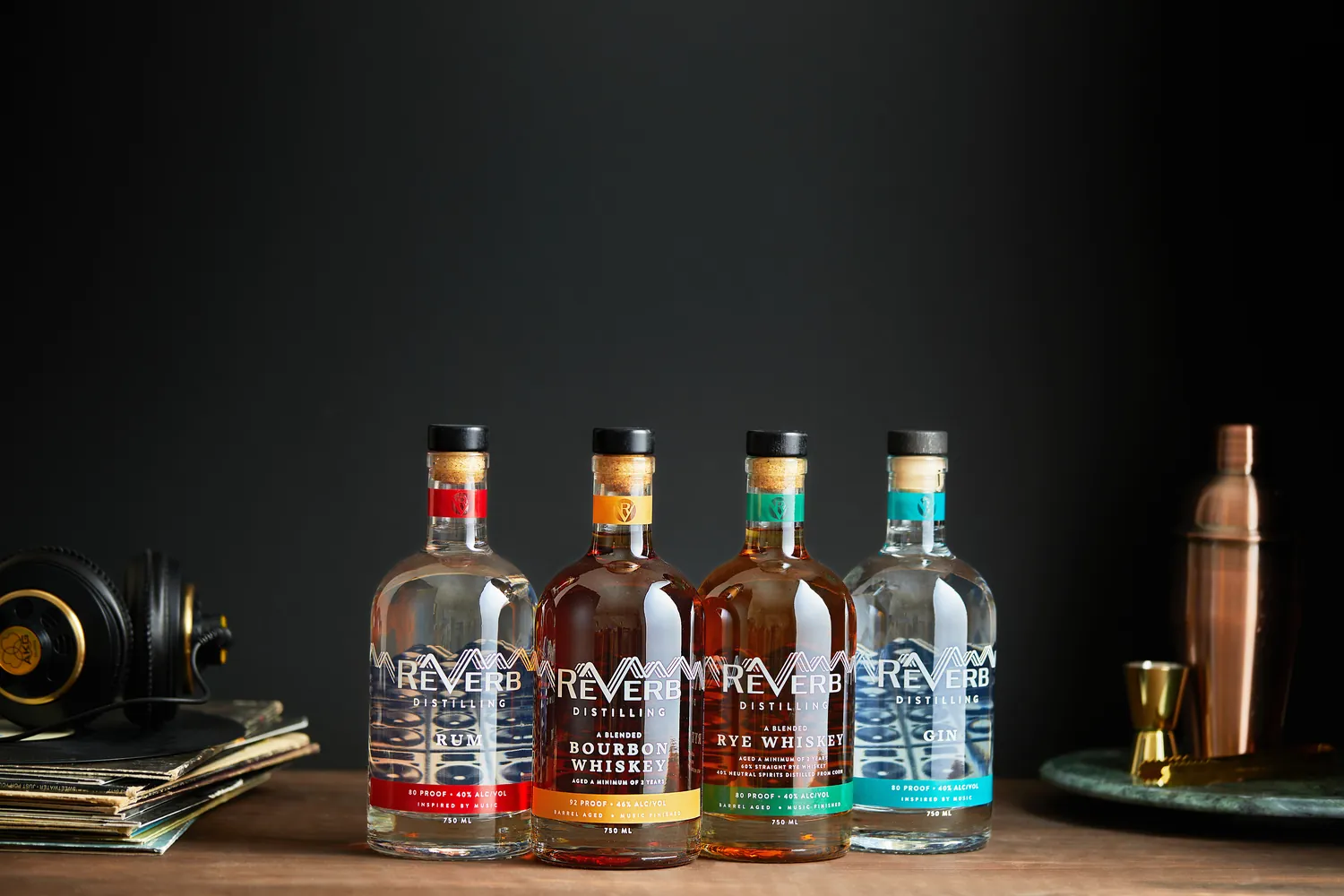 ReVerb Rum, Bourbon Whiskey, Rye Whiskey and Gin standing side by side on a wooden table, with headphones and records at the left and a bar shaker at the right